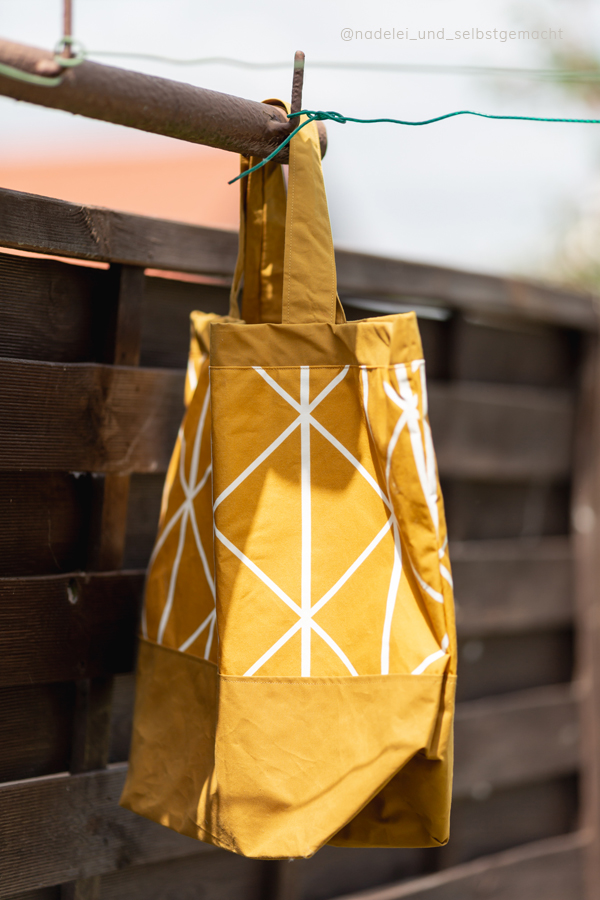Ockerfarbene Tasche mit Boden an einem Metallrohr hängend. Der Hintergrund ist dunkles Holz. Die Tasche ist selber genäht nach dem Schnittmuster Carry&Go