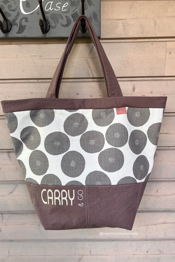 „Selbstgenähte Shopper-Tasche im Carry-&-Go-Stil, hängend an einer Garderobenleiste vor heller Holzwand. Der Hauptteil zeigt ein grafisches Stoffmuster mit großen grauen Kreisen auf weißem Grund, kombiniert mit taupefarbenen Akzenten an Boden, oberer Blende und langen Tragehenkeln. Vorne links ist der Schriftzug ‚CARRY & GO‘ zu sehen."