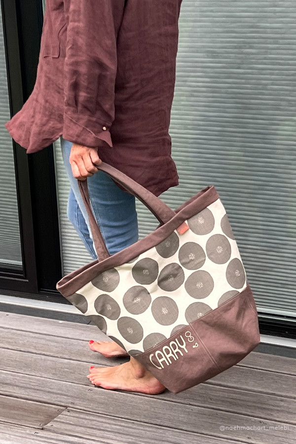 Einkaufstasche genäht nach Schnittmuster Carry&Go in Taupe mit grafischem Dessin im Mittelteil. Getragen wird die Tasche von einer Frau mit braunem Leinenhemd. Sie ist barfuss auf einer Holzterrasse.