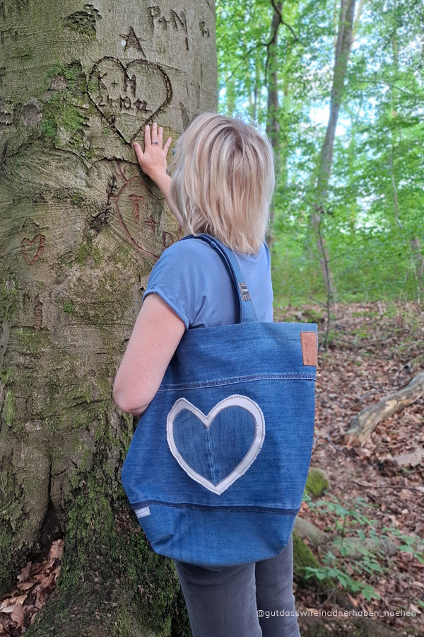 selbstgenähte legere Stofftasche aus alten Jeans mit Herzapllikation. Eine blonde Frau trägt sie über der Schulter. Sie steht vor einem Baum und bewundert ein geschnitztes Herz.
