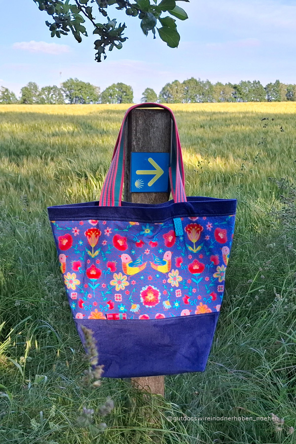 Große selbstgenähte Stofftasche in kräftigen Blautönen mit floral gemusterten Mittelteil. Location: Am Rande eines Kornfeldes am Holzpfahl hängend