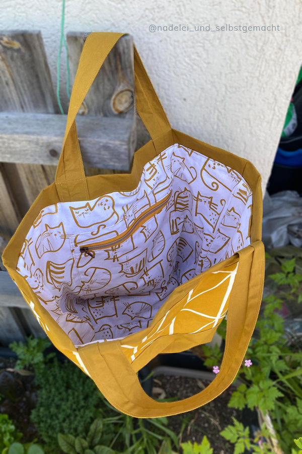 Innenansicht einer selbgenähten Stofftasche aus ockerfarbenem Oilskin. Das Futter ist weiß mit farblich zum Aussenstoff passenden gezeichneten Katzen. Sichtbar ist auch ein Reißverschlussfach.