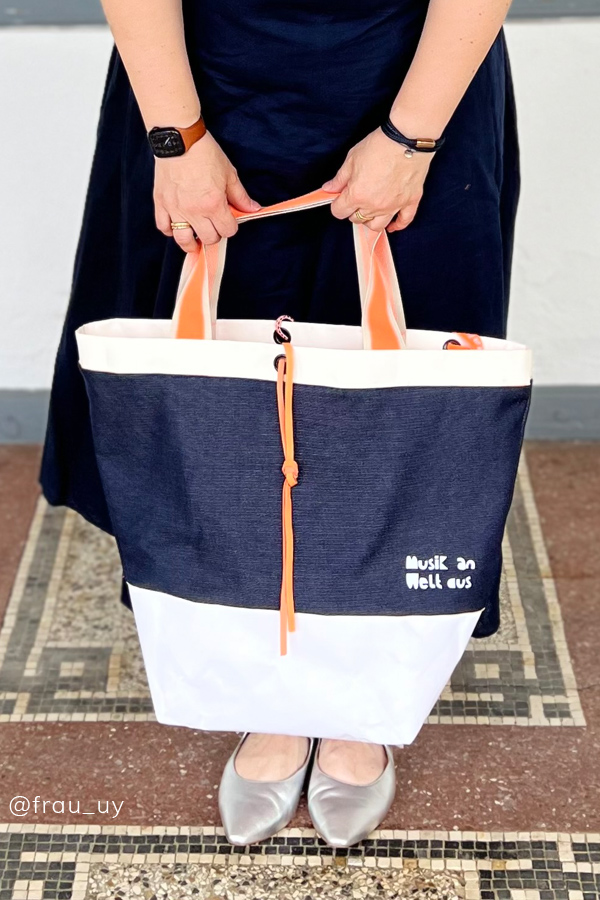 Selbstgenähte Tasche mit Boden in Marineblau und Weiß mit orangefarbenen Henkeln und Stickschrift ‚Musik an, Welt aus', gehalten von einer Frau in dunklem Kleid