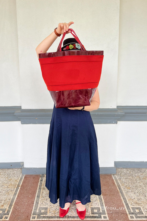 Frau in dunkelblauem Sommerkleid und roten Schuhen hält eine selbstgenähte Shopper-Tasche aus rotem Stoff mit Lederdetails in Rot- und Bordeauxtönen
